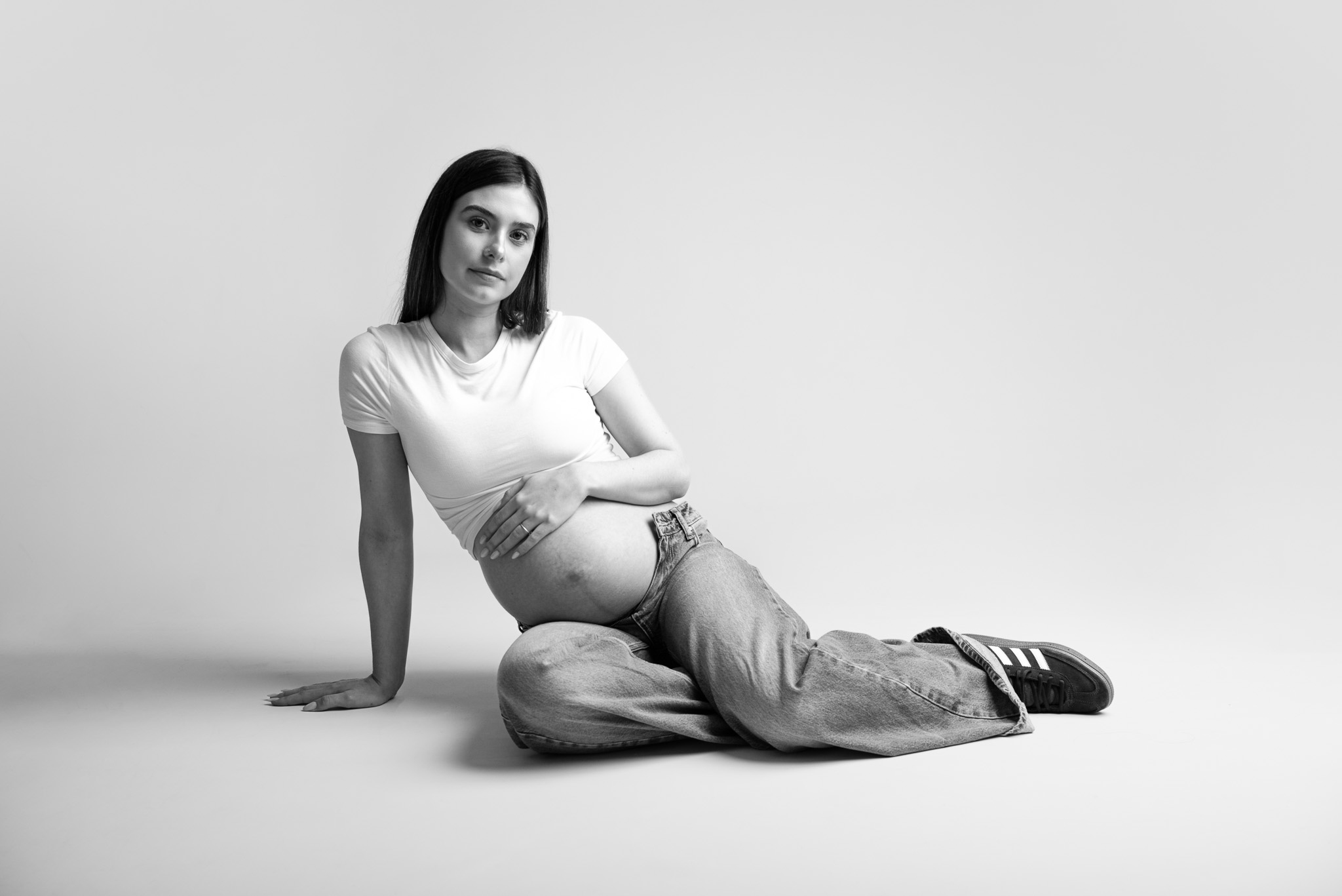 Photo de grossesse en noir et blanc réalisée en studio par une photographe maternité, montrant une femme enceinte assise au sol, pose naturelle et fond neutre.
